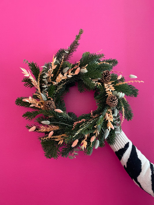 Couronne de noel vert sauge et terracotta sur fond rose fushia tenu par une main au pull zébré