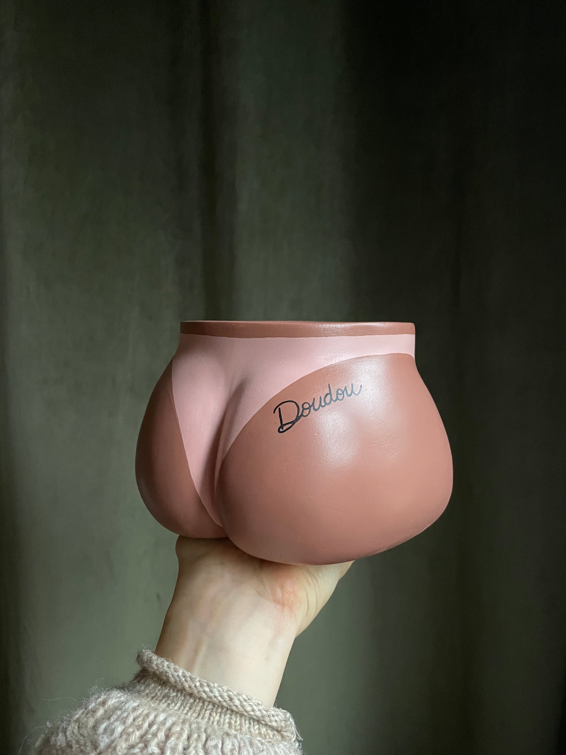 Cache-pot Fessier côté fesses tenu à la main par le dessous, avec une marque de bronzage de bikini  et un tatouage personnalisé avec le prénom "Doudou" écrit en noir en écriture cursive sur la fesse droite, sur fond vert kaki, objet déco original corps féminin fait main en résine écologique (Jesmonite) par Maison Tessier.