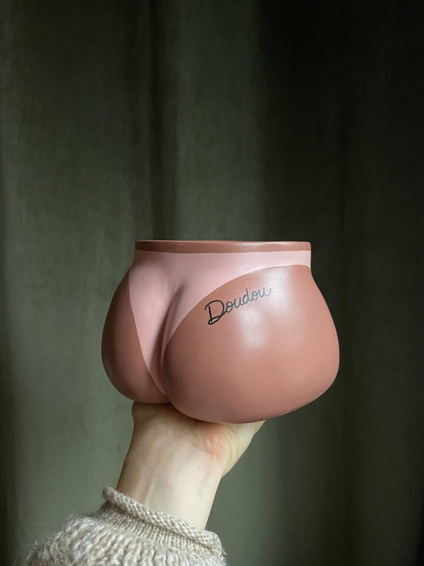 Cache-pot Fessier côté fesses tenu à la main par le dessous, avec une marque de bronzage de bikini  et un tatouage personnalisé avec le prénom "Doudou" écrit en noir en écriture cursive sur la fesse droite, sur fond vert kaki, objet déco original corps féminin fait main en résine écologique (Jesmonite) par Maison Tessier.