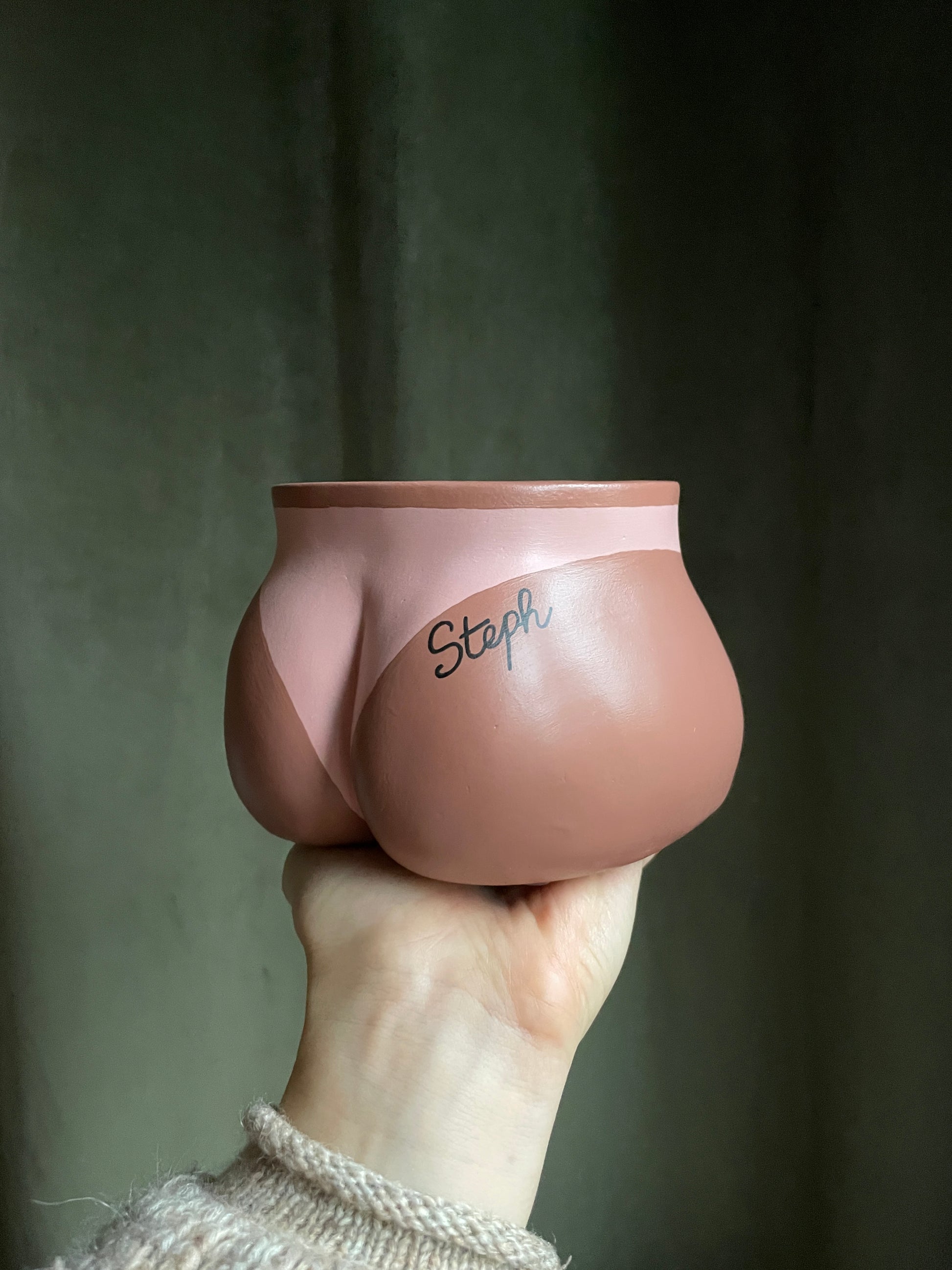 Cache-pot Fessier côté fesses tenu à la main par le dessous, avec une marque de bronzage de bikini  et un tatouage personnalisé avec le prénom "Steph" écrit en noir en écriture cursive sur la fesse droite, sur fond vert olive, objet déco original corps féminin fait main en résine écologique (Jesmonite) par Maison Tessier.