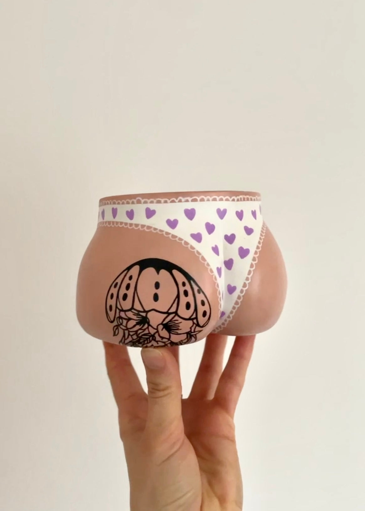 Cache-pot Fessier Culotté Coeurs côté fesses coloris crème tenu à la main sur un fond crème, avec une culotte blanche à dentelle et petits cœurs parme, et un tatouage personnalisé sur la fesse gauche, reprenant celui d’une cliente, objet déco original corps féminin fait main en résine écologique par Maison Tessier. 