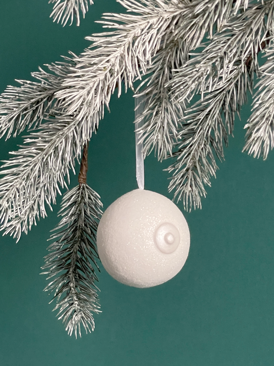 Le Saint des Seins en forme de sein avec paillettes blanches couleur neige et le téton blanc nacré accroché sur une branche de sapin, sur fond vert émeraude.