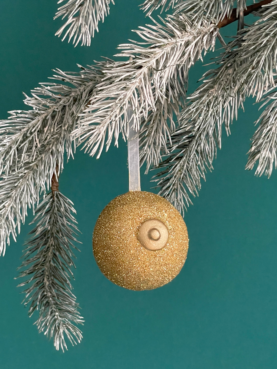 Le Saint des Seins en forme de sein avec paillettes or et le téton or accroché sur une branche de sapin, sur fond vert émeraude.