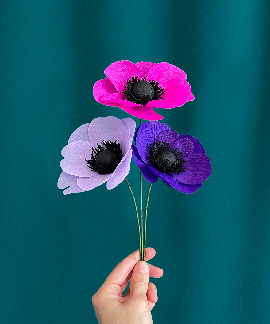 trois fleurs en papier anémones de couleur fushia, parme et violet, tenues dans une main sur un fond couleur vert émeraude