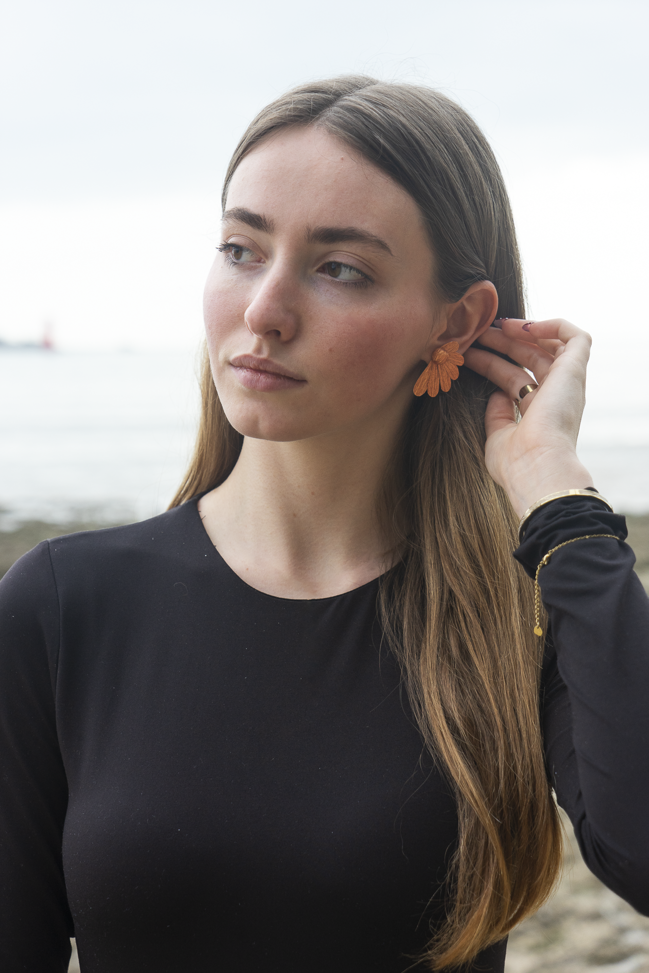 Femme portant une boucle d’oreille Puce Pâquerettes orange clair, coeur cuivre en papier crépon fait main au bord de la mer, bijoux floraux artisanaux par Maison Tessier.