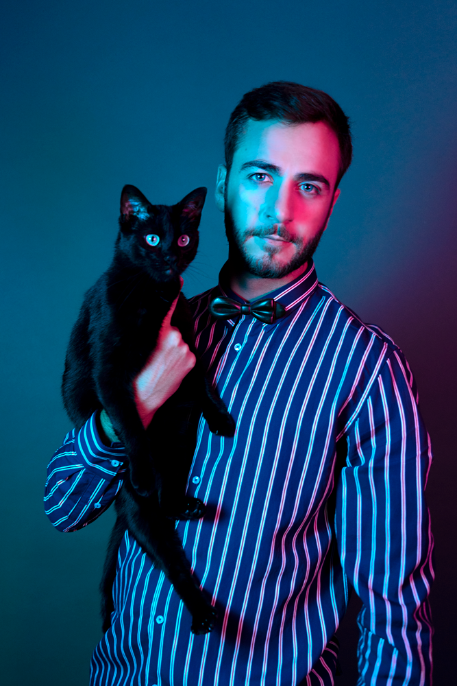 Homme avec portant un chat noir, il est habillé avec une chemise à rayure bleue et blanche et un noeud papillon en bois Lysandra Dalbergia Maison Tessier