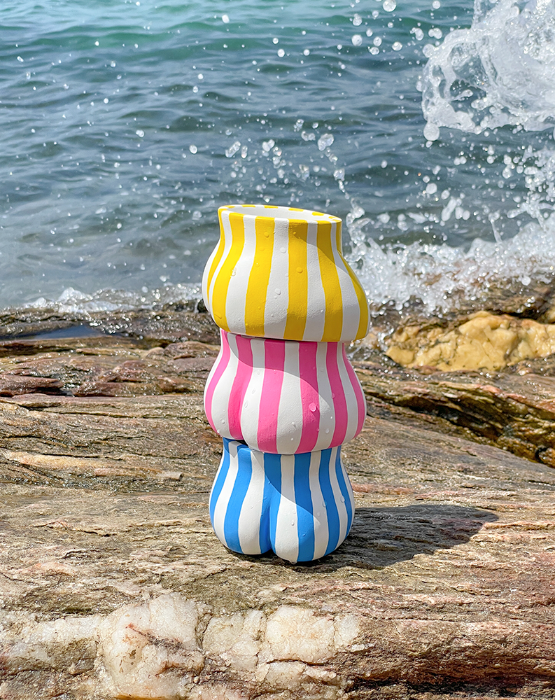 3 Fessiers Cabanon, bleu, rose, jaune, empilés sur un rocher face à la mer avec une vague en fond