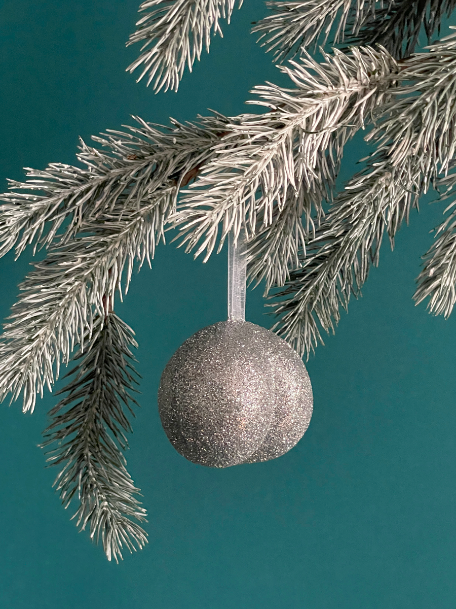 Le Boule de Noël en forme de fesses avec des paillettes Argent accroché sur une branche de sapin, sur fond vert émeraude.