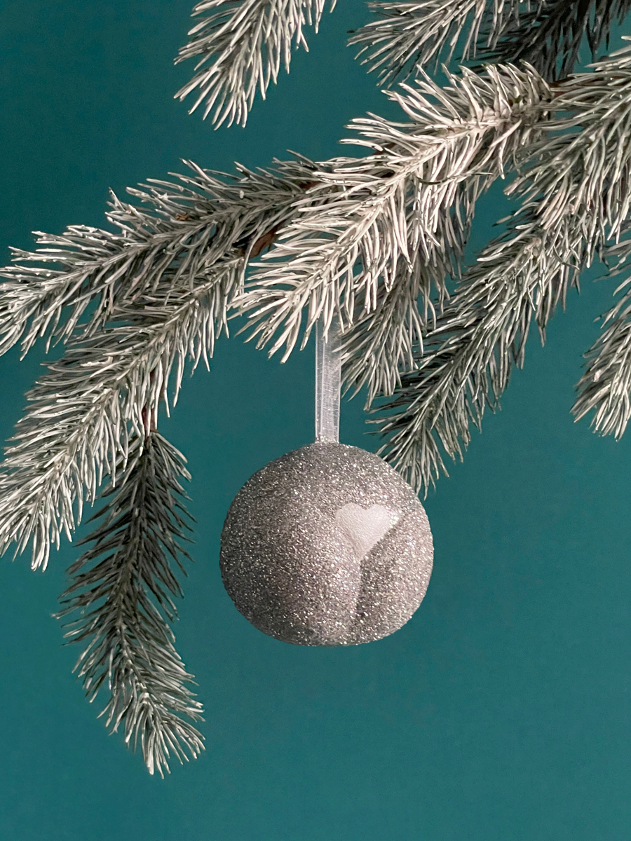 Le Boule de Noël en forme de pubis avec paillettes Argent et petit coeur Argent nacré, accroché sur une branche de sapin sur fond vert émeraude.