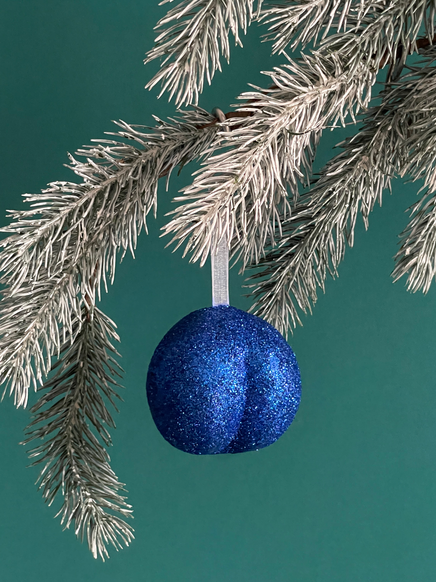 Le Boule de Noël en forme de fesses avec des paillettes Bleues accroché sur une branche de sapin, sur fond vert émeraude.