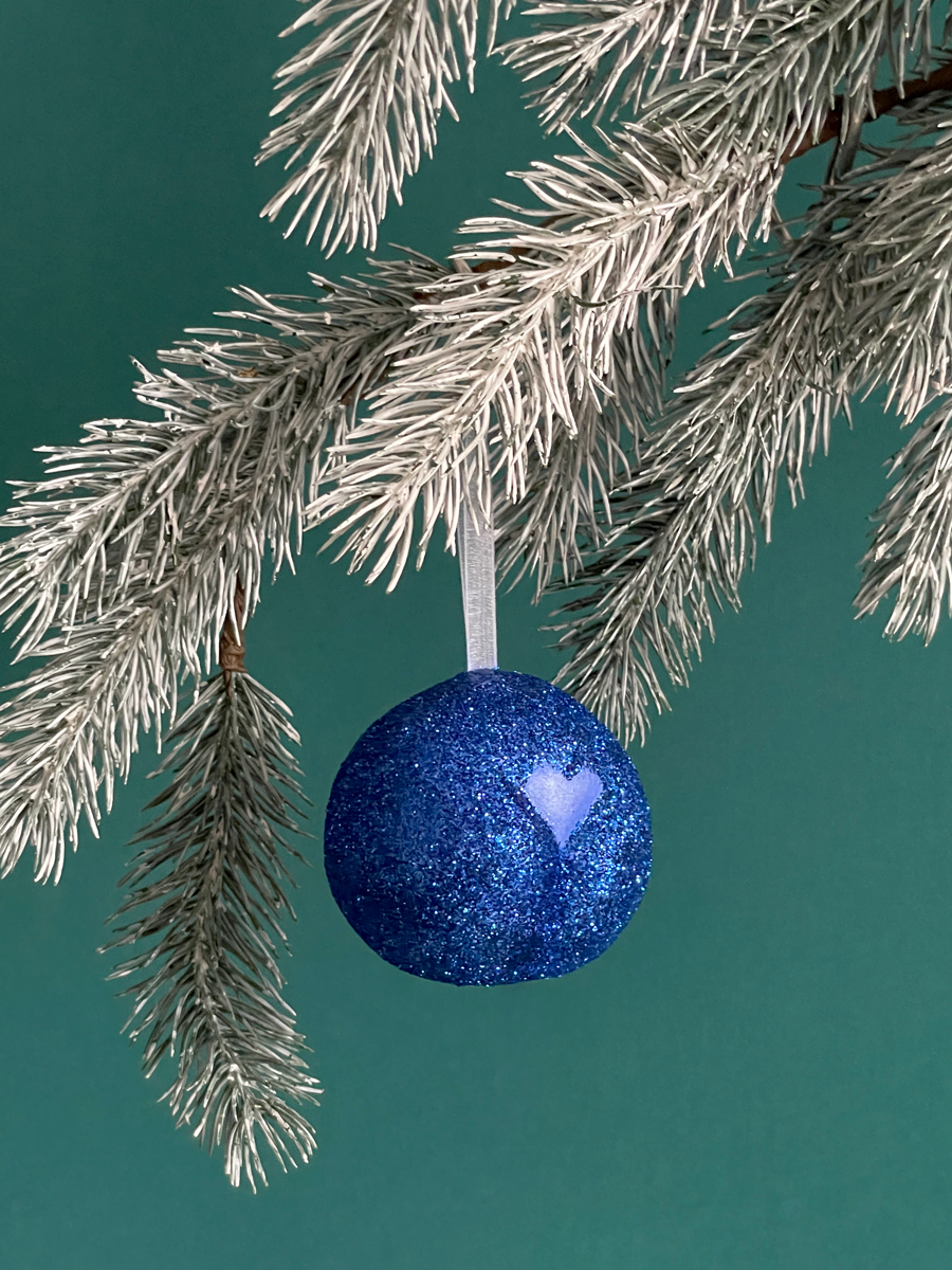 Le Boule de Noël en forme de pubis avec paillettes Bleues et petit coeur Bleu nacré, accroché sur une branche de sapin sur fond vert émeraude.