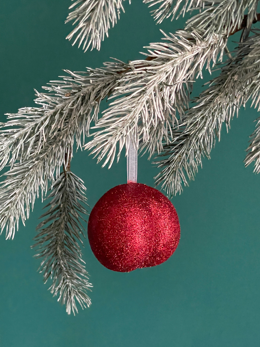Le Boule de Noël en forme de fesses avec des paillettes rouges accroché sur une branche de sapin, sur fond vert émeraude.