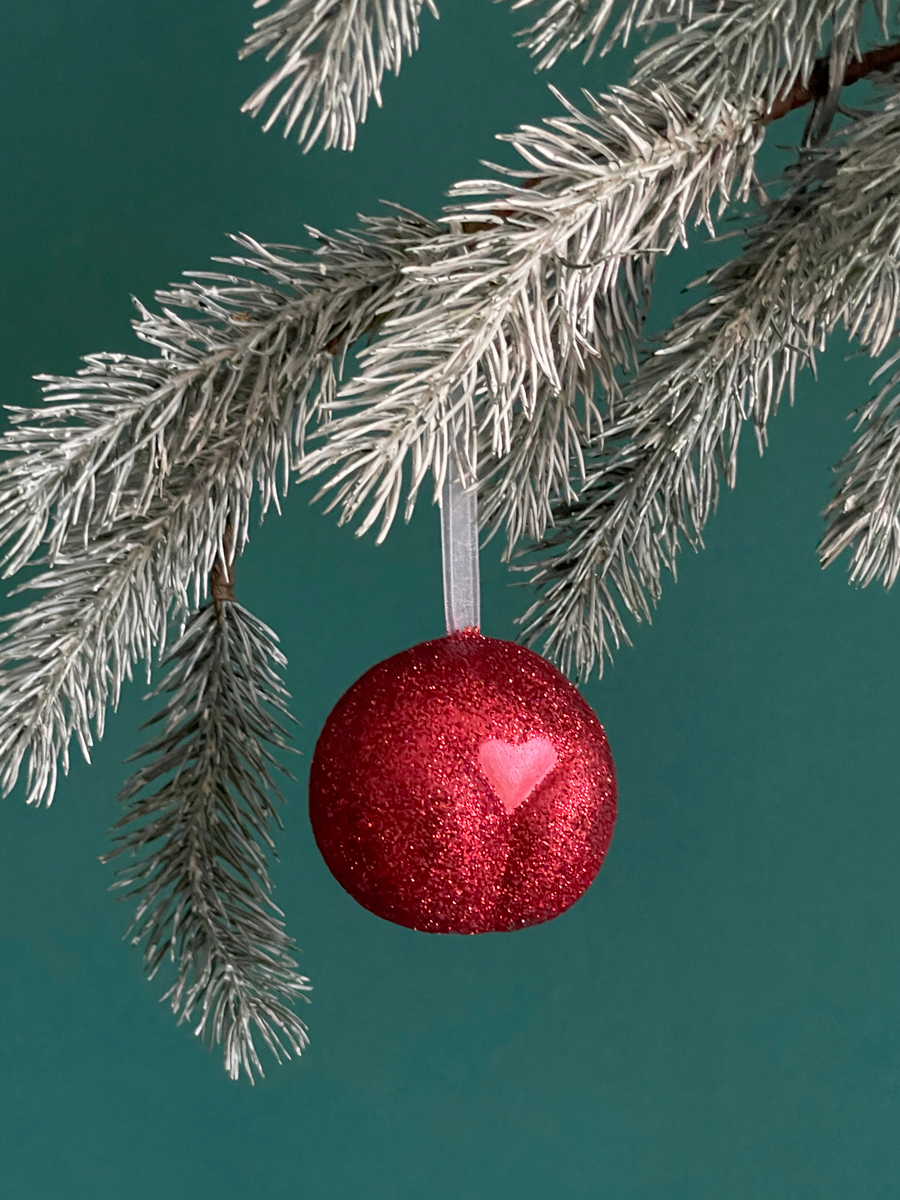 Le Boule de Noël en forme de pubis avec paillettes rouges et petit coeur rouge nacré, accroché sur une branche de sapin sur fond vert émeraude.