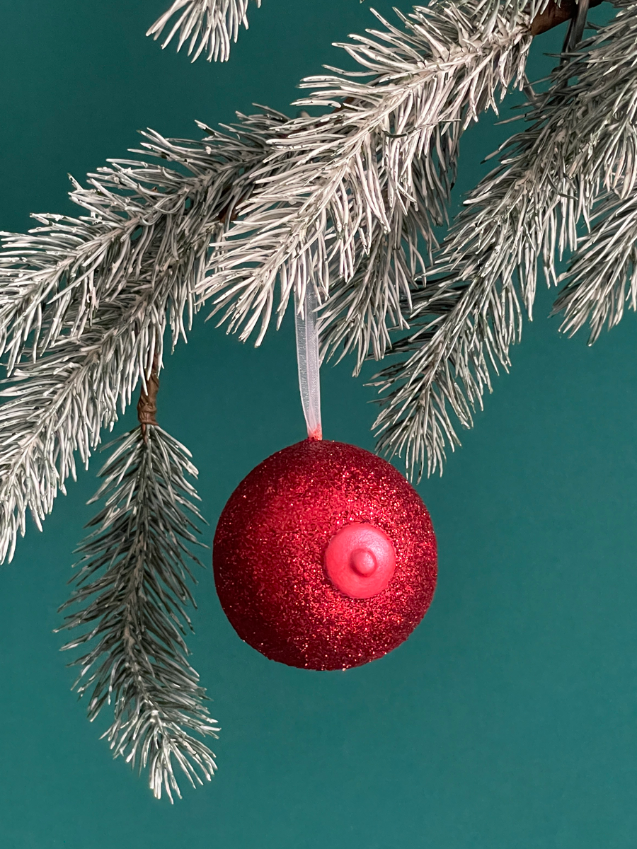 Le Saint des Seins en forme de sein avec paillettes rouges et le téton rouge nacré accroché sur une branche de sapin, sur fond vert émeraude.