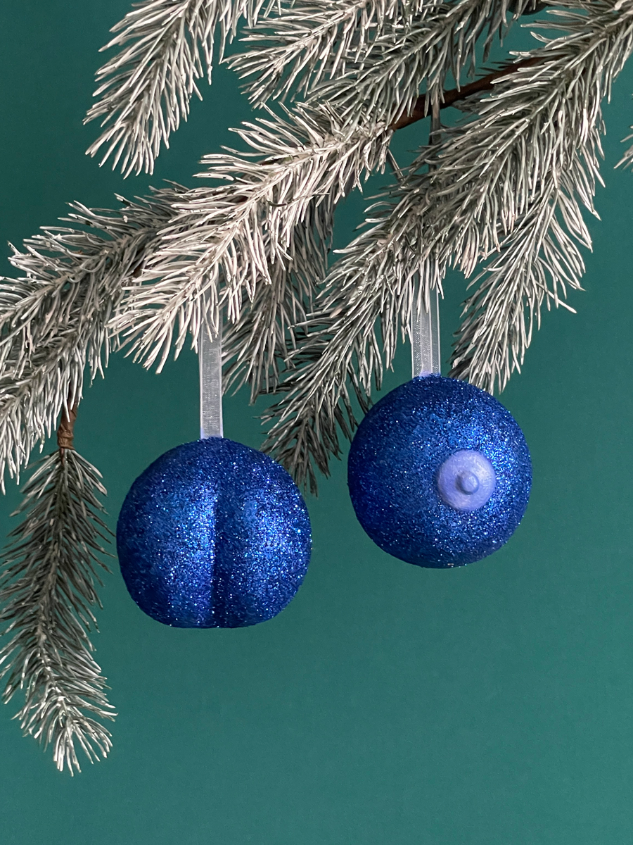 Paire de boules de Noël Maison Tessier, avec le Boule de Noël en forme de pubis avec paillettes Bleues et petit coeur bleu nacré et du Saint des Seins avec des paillettes Bleues et le téton bleu nacré accrochés sur une branche de sapin, sur fond vert émeraude.