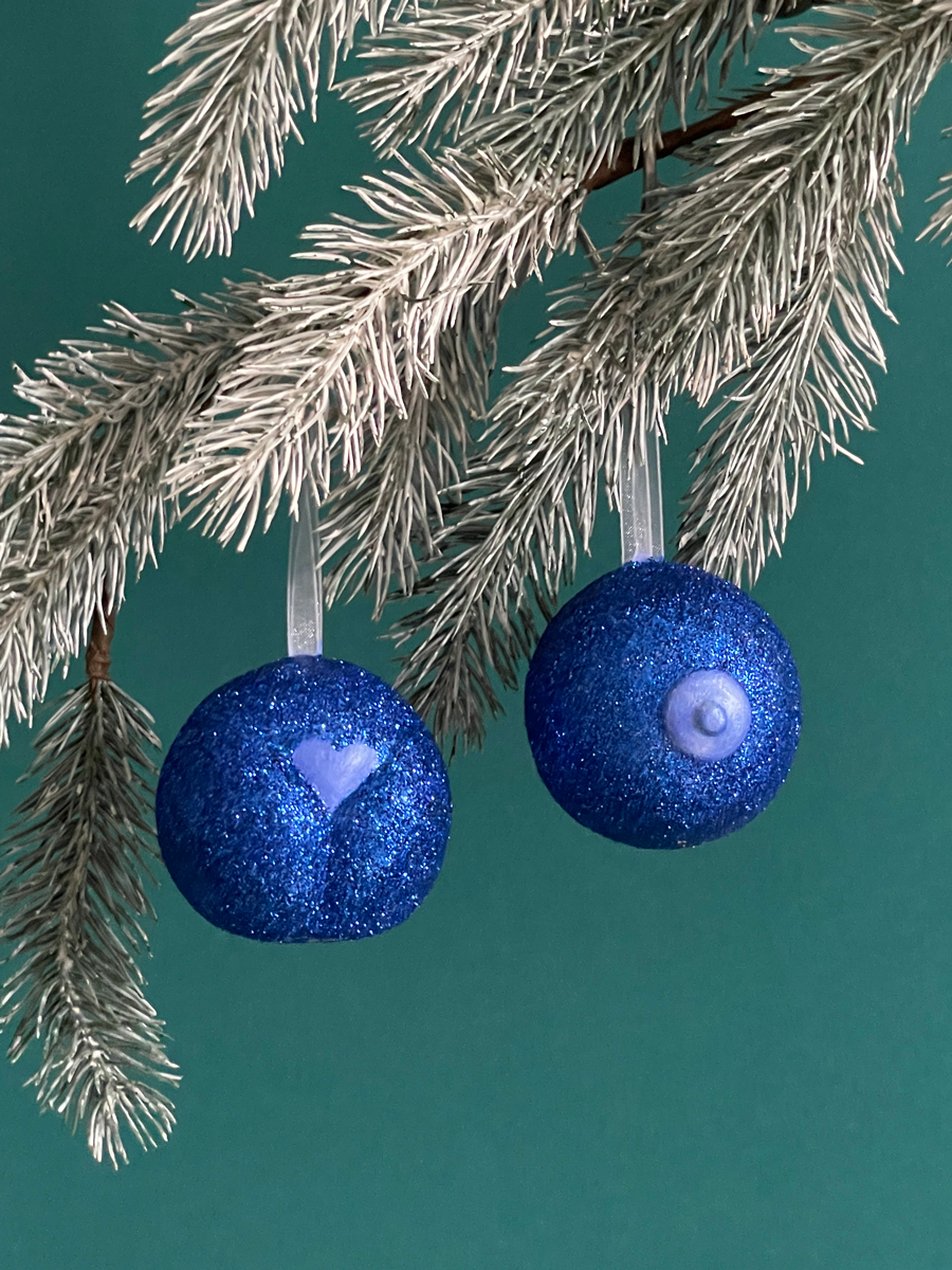 Paire de boules de Noël Maison Tessier, avec le Boule de Noël en forme fesses avec des paillettes Bleues et du Saint des Seins avec des paillettes Bleues et le téton bleu nacré accrochés sur une branche de sapin, sur fond vert émeraude.