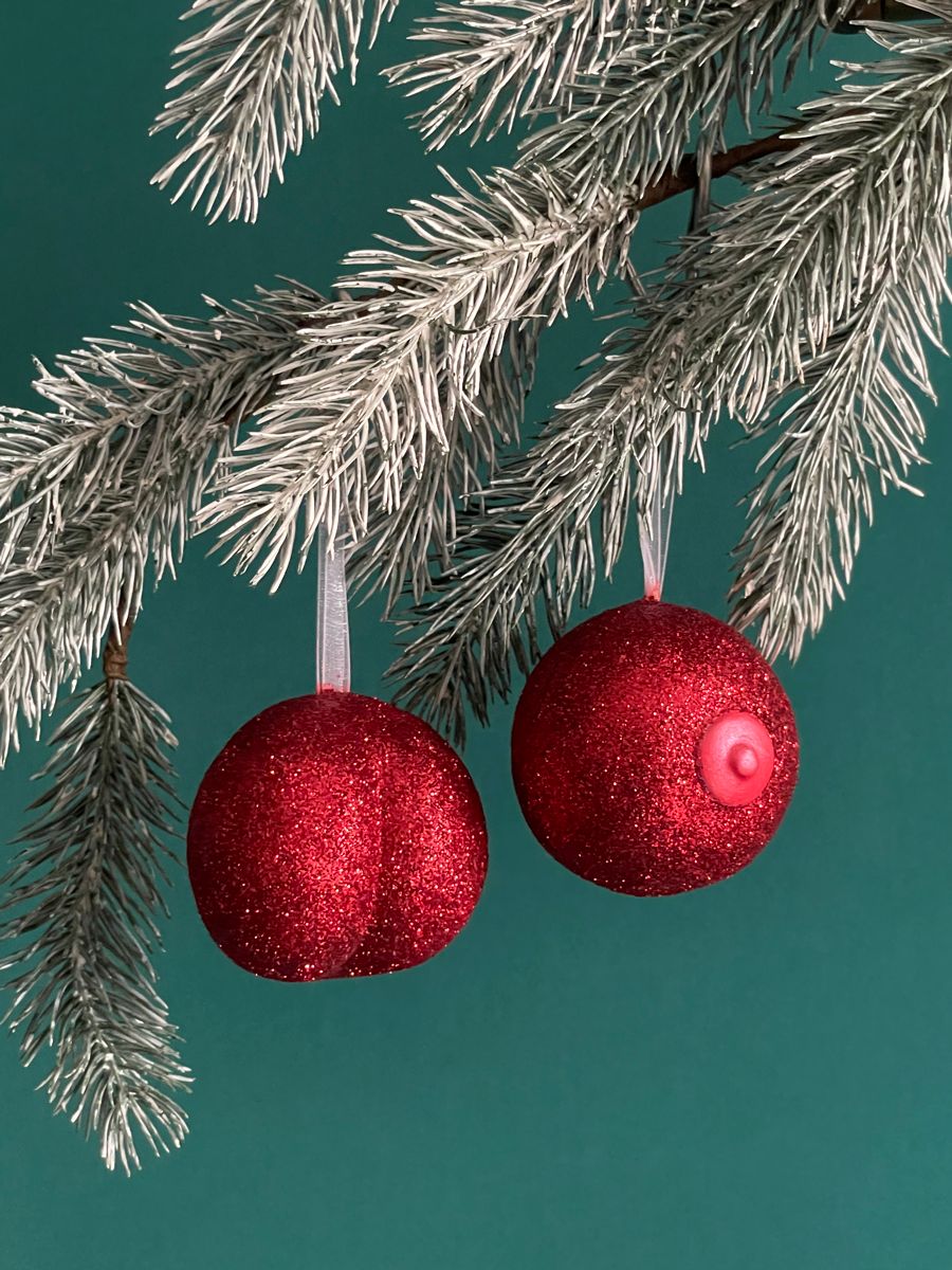 Paire de boules de Noël Maison Tessier, avec le Boule de Noël en forme fesses avec des paillettes rouges et du Saint des Seins avec des paillettes rouges et le téton rouge nacré accrochés sur une branche de sapin, sur fond vert émeraude.