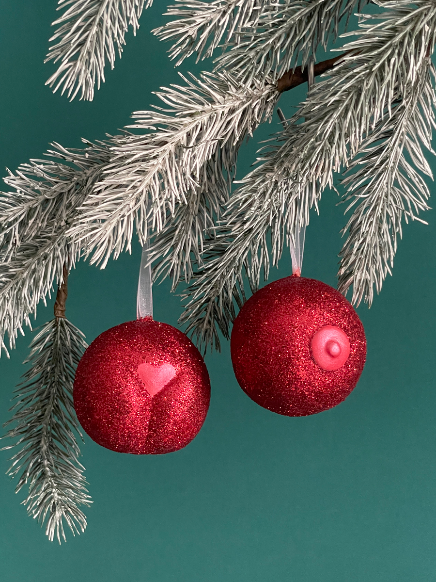 Paire de boules de Noël Maison Tessier, avec le Boule de Noël en forme de pubis avec paillettes rouges et petit coeur rouge nacré et du Saint des Seins avec des paillettes rouges et le téton rouge nacré accrochés sur une branche de sapin, sur fond vert émeraude.