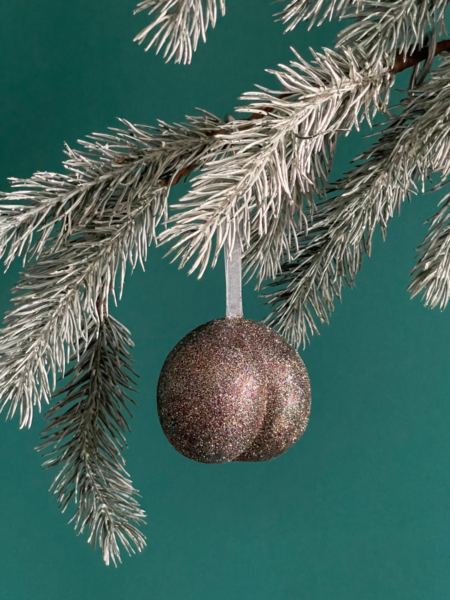 Le Boule de Noël en forme de fesses avec des paillettes Bronze accroché sur une branche de sapin, sur fond vert émeraude.