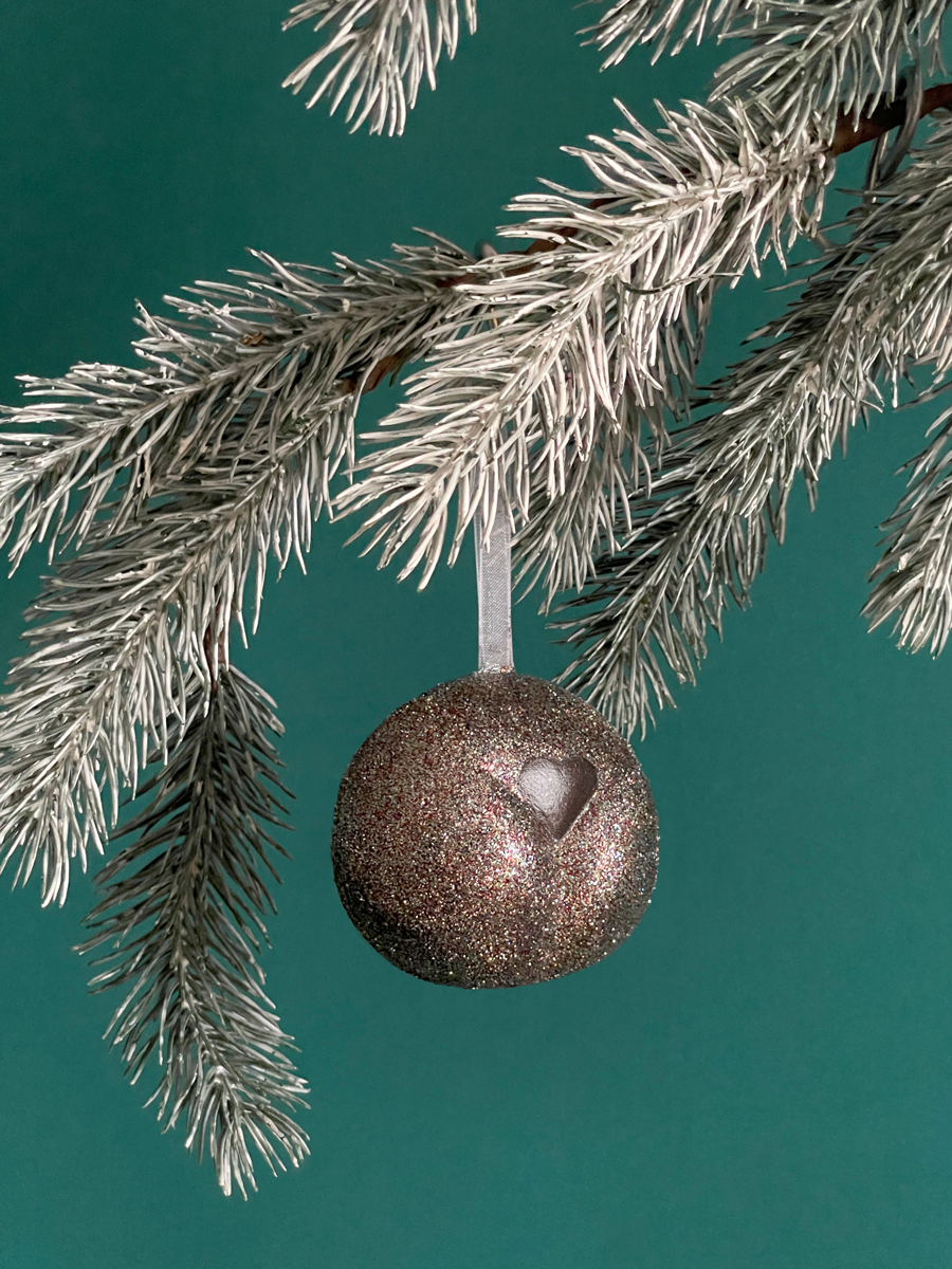 Le Boule de Noël en forme de pubis avec paillettes Bronze et petit coeur Bronze nacré, accroché sur une branche de sapin sur fond vert émeraude.