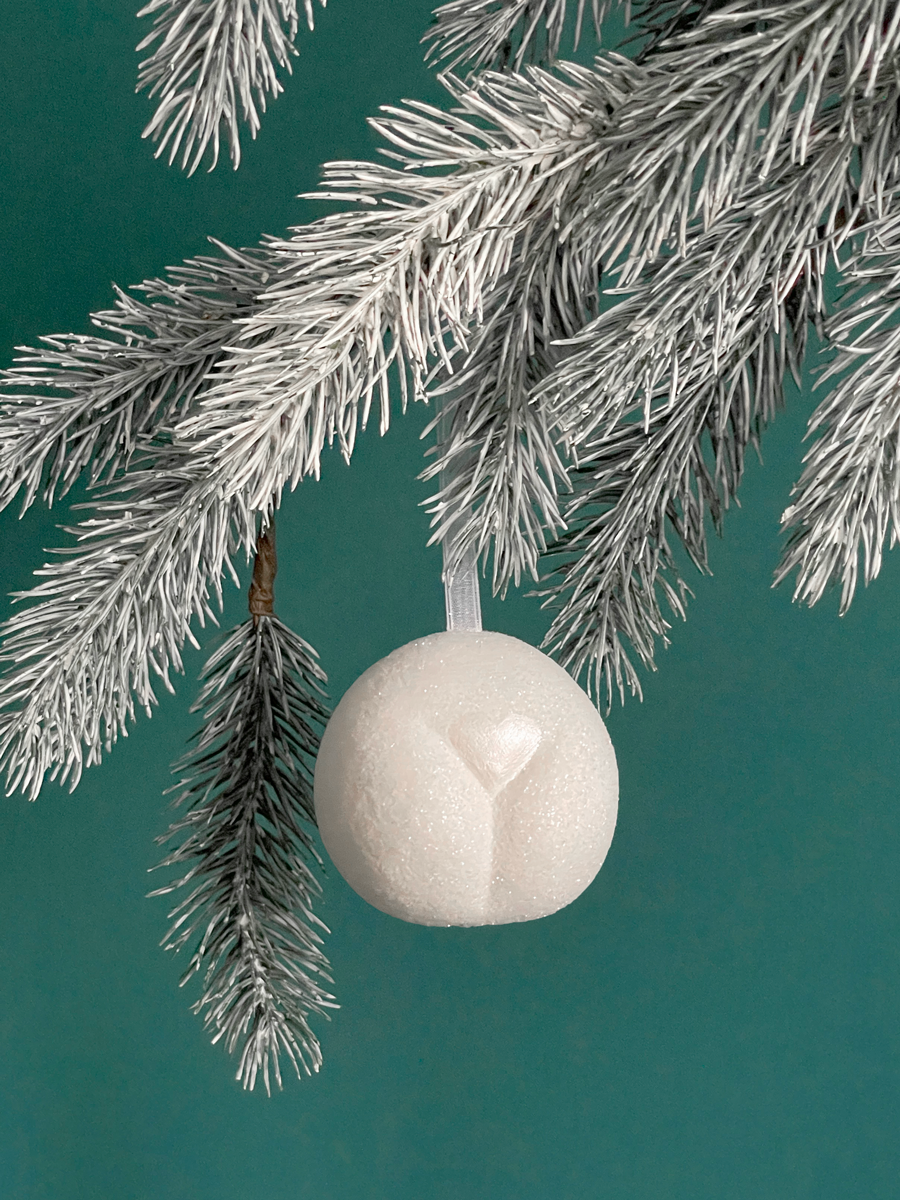Le Boule de Noël en forme de pubis avec paillettes Neige et petit coeur Neige nacré, accroché sur une branche de sapin sur fond vert émeraude.