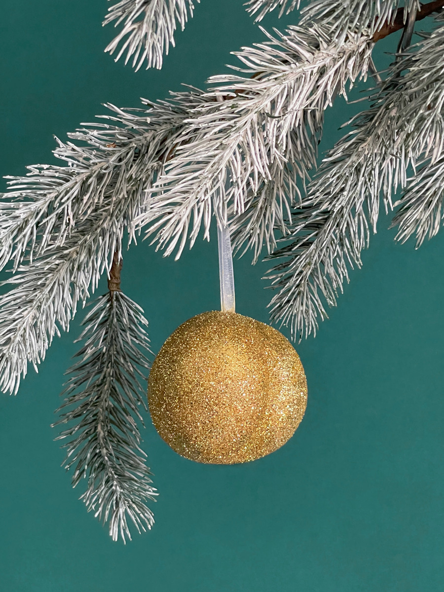 Le Boule de Noël en forme de fesses avec des paillettes Or accroché sur une branche de sapin, sur fond vert émeraude.