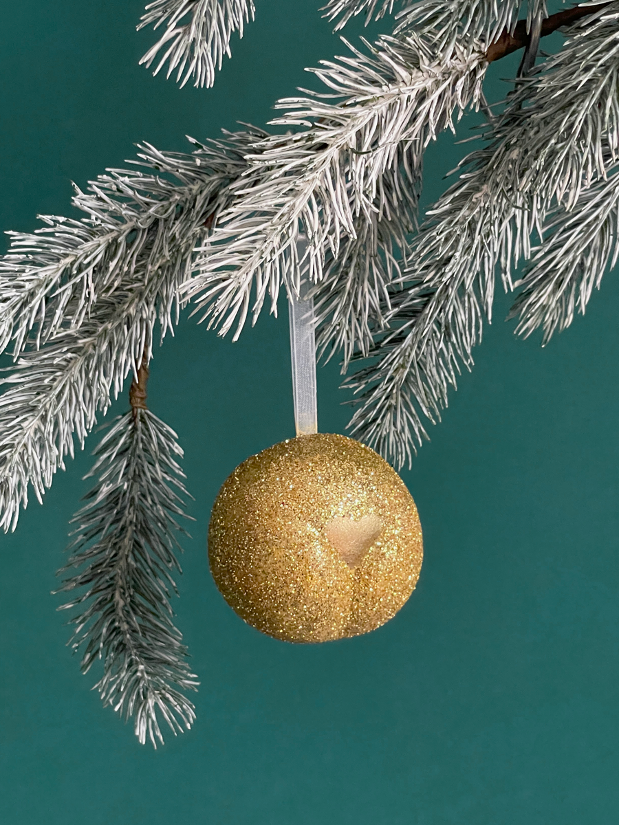 Le Boule de Noël en forme de pubis avec paillettes or et petit coeur or nacré, accroché sur une branche de sapin sur fond vert émeraude.