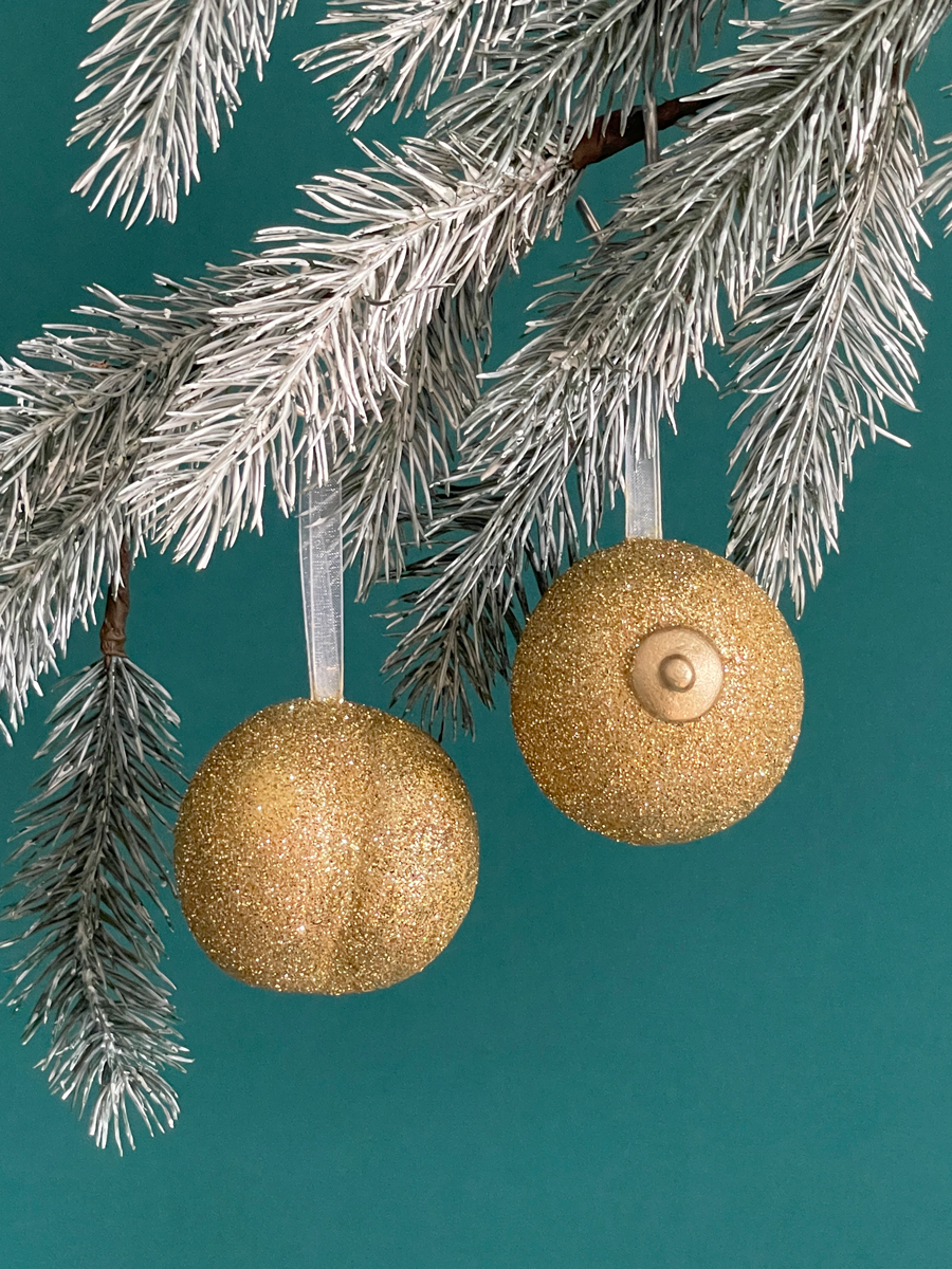 Paire de boules de Noël Maison Tessier, avec le Boule de Noël en forme fesses avec des paillettes or et du Saint des Seins avec des paillettes or et le téton or accrochés sur une branche de sapin, sur fond vert émeraude.