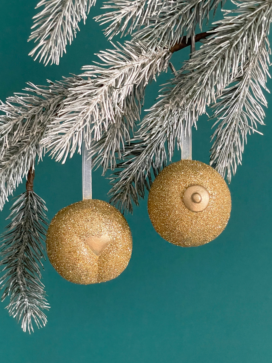 Paire de boules de Noël Maison Tessier, avec le Boule de Noël en forme de pubis avec paillettes or et petit coeur or et du Saint des Seins avec des paillettes or et le téton or accrochés sur une branche de sapin, sur fond vert émeraude.