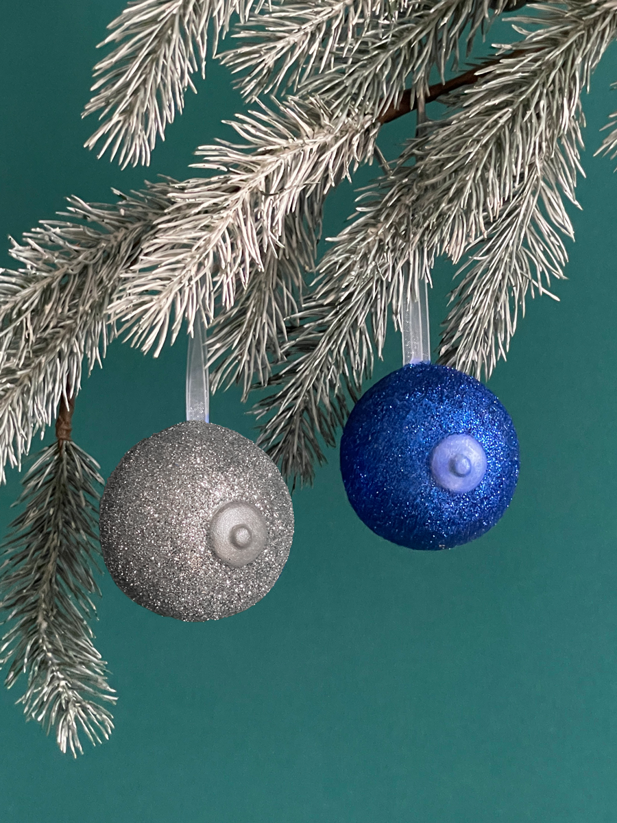 Paire de boules de Noël Maison Tessier, avec le Saint des Seins en forme de sein avec paillettes argent et le téton argent et l’autre Sein avec des paillettes bleu et le téton bleu nacré accrochés sur une branche de sapin, sur fond vert émeraude.