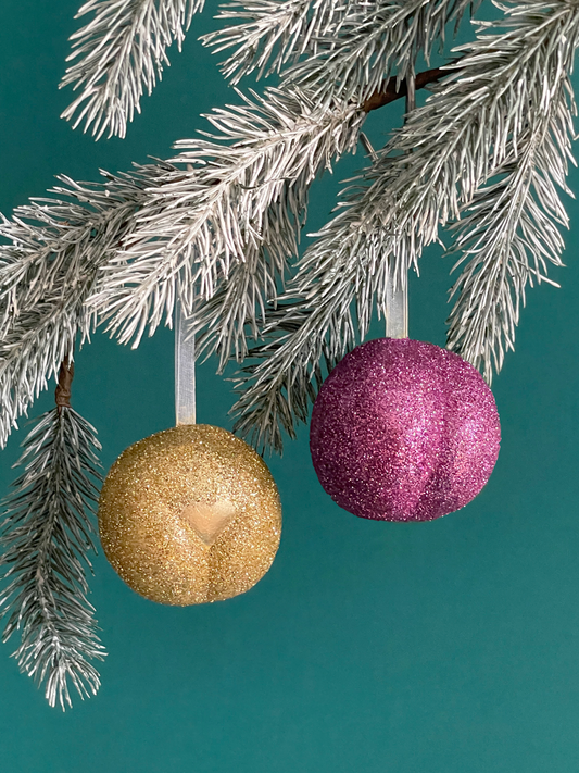 Paire de boules de Noël Maison Tessier, avec le Boule de Noël en forme de pubis avec paillettes d'or et petit coeur or et de fesses avec des paillettes roses accrochés sur une branche de sapin, sur fond vert émeraude.