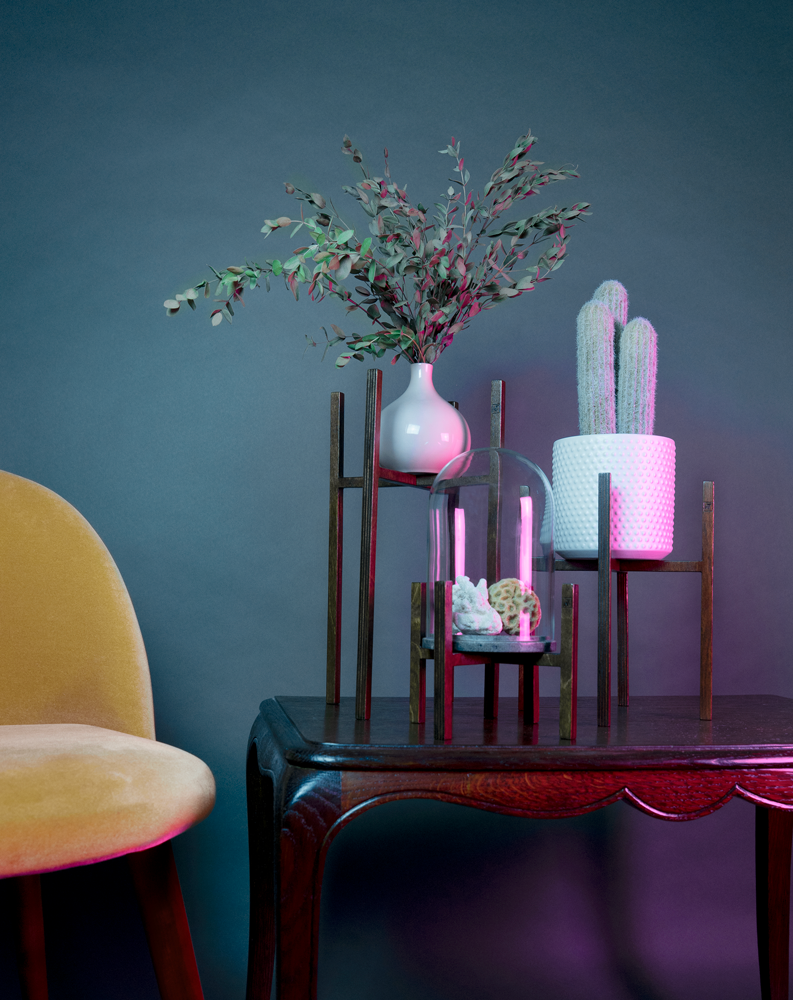 Trois piédestaux, de la série ITUS de la marque Maison Tessier, portent chacun un objet de déco, se tiennent sur une table en bois, derrière un fond bleu avec des reflets de lumière rose, à côté se trouve un fauteuil en velours jaune