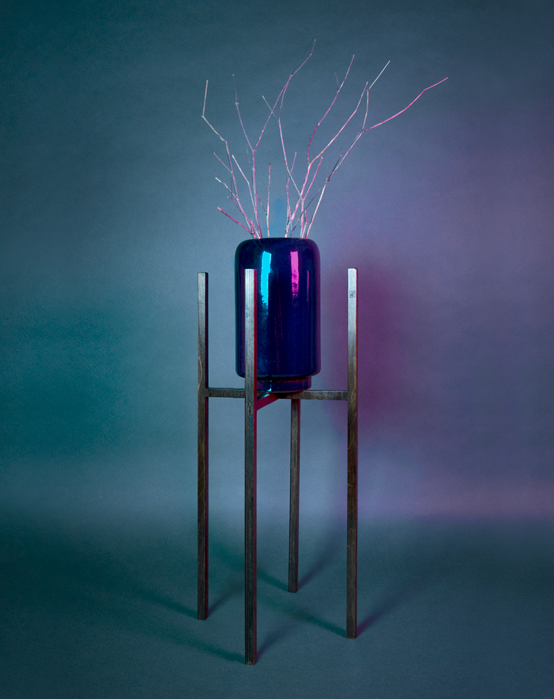 Pot en céramique émaillé bleu foncé avec des branches est soutenu par un piédestal ITUS INGENTIS de la marque Maison Tessier derrière un fond bleu avec des reflets de lumière rose