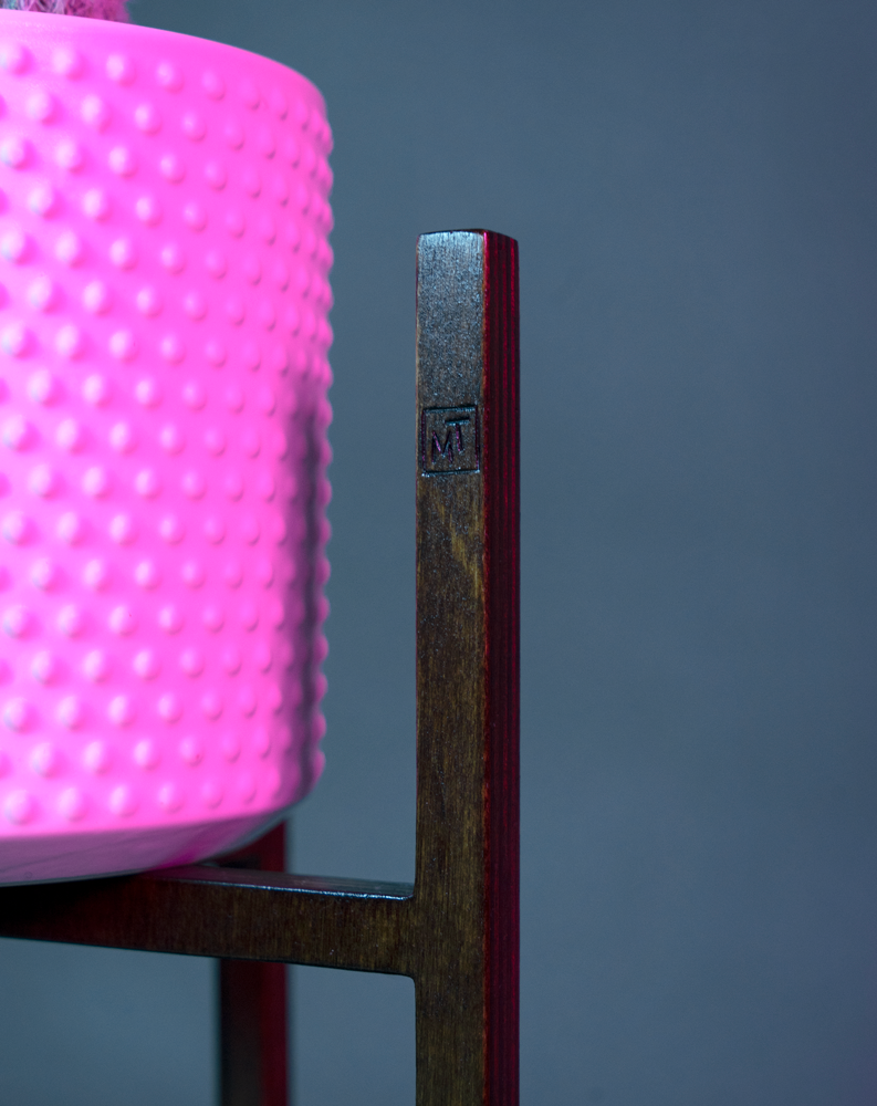 Pot en céramique émaillé blanc, contenant des cactus, est soutenu par un piédestal ITUS MAXIMA de la marque Maison Tessier derrière un fond bleu avec des reflets de lumière rose, zoom sur la signature