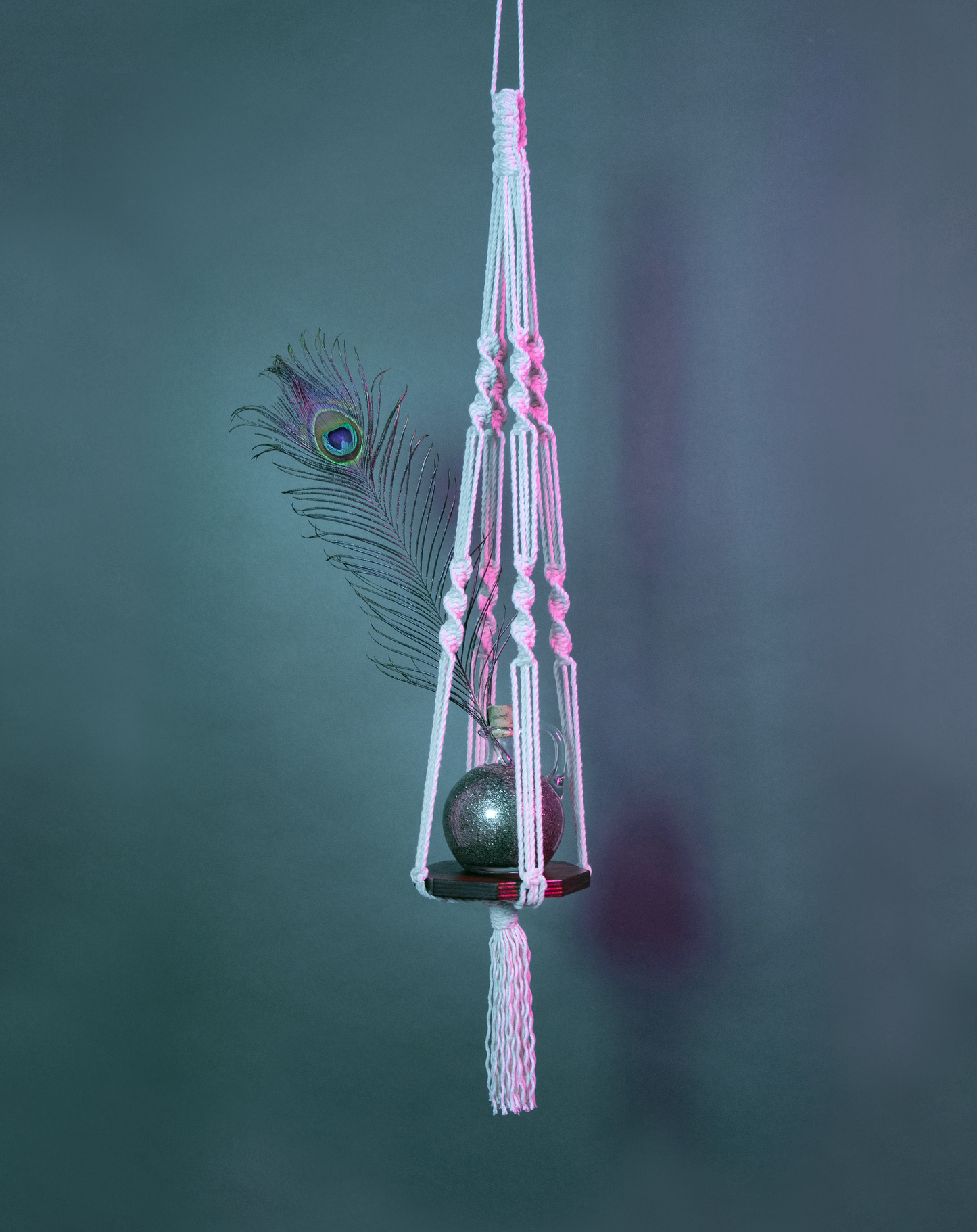 Une suspension en macramé de coton et plateau en bois PLANIUM MINIMA, de la marque Maison Tessier, soutient un pot de sable et une plume de paon, derrière un fond bleu avec des reflets de lumière rose
