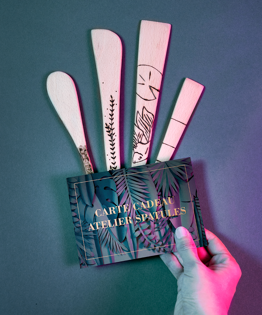 Carte cadeau atelier spatules Maison Tessier avec 4 spatules différentes posées derrière la carte tenue par une main